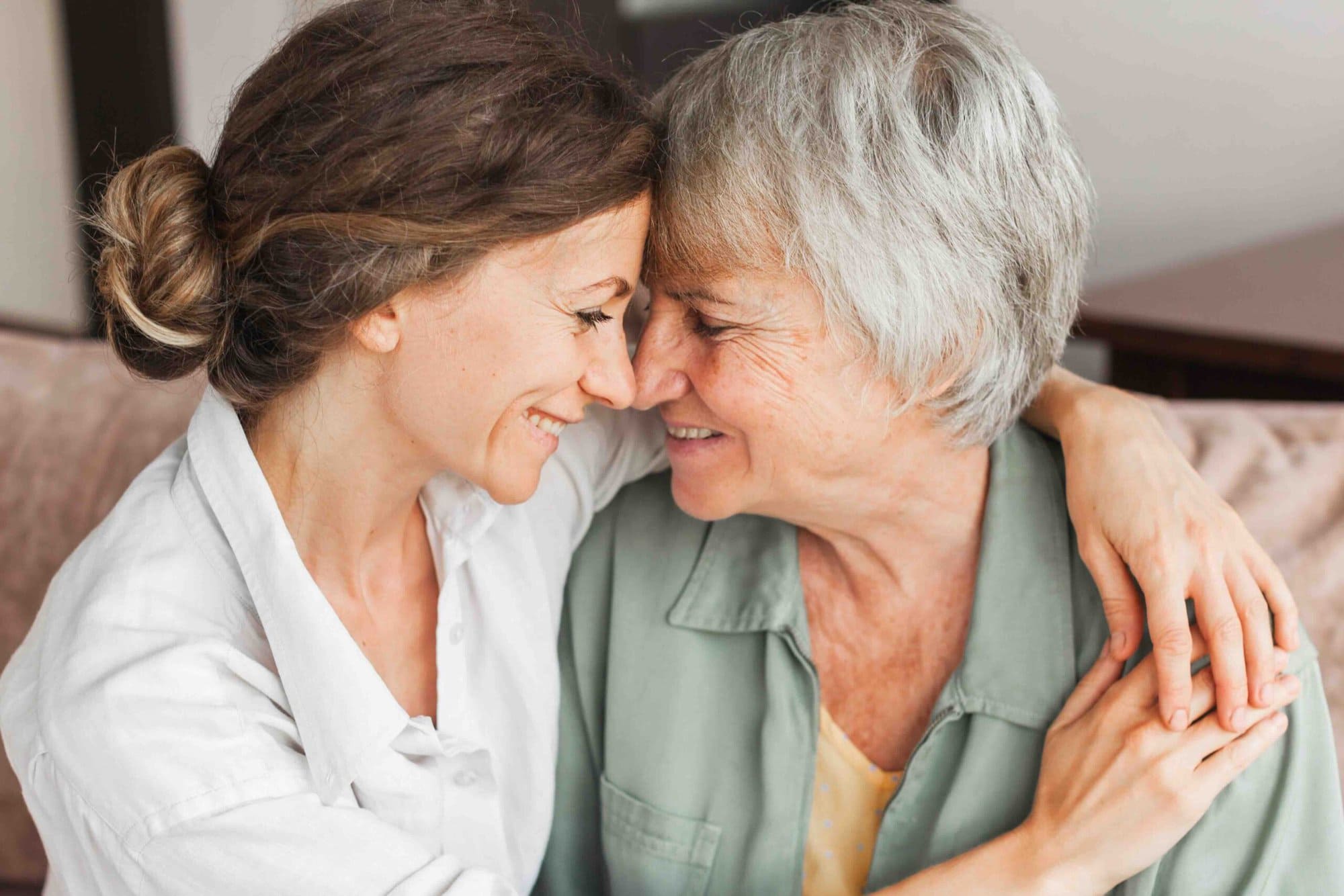 A woman and her mother resting their foreheads together, laughing at home